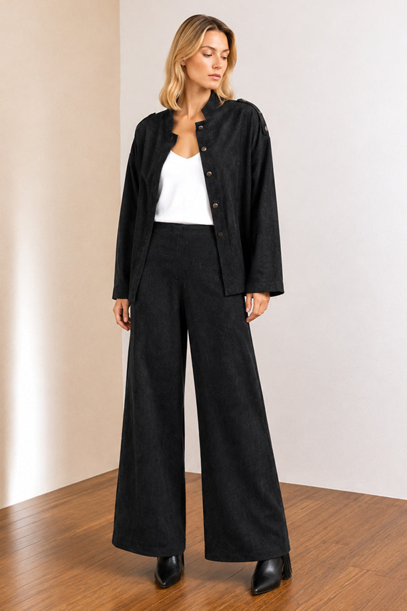 A woman modeling a black textured jacket and matching wide-leg trousers with a white top, standing in a minimalist room with beige walls and a wooden floor.
