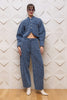 A model stands against a textured beige wall wearing a matching blue tweed cropped jacket and wide-leg trousers set, paired with white pointed-toe heels.